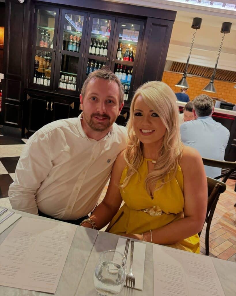 About Us 2 Madison Cole sitting beside her husband at a cozy restaurant, wearing a yellow dress and smiling warmly across the table.