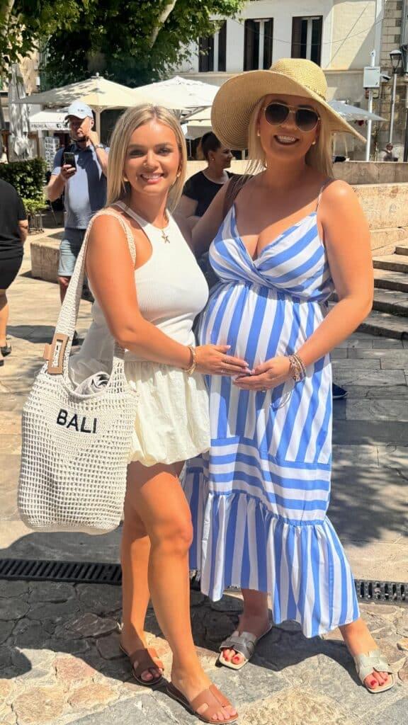 About Us 1 Madison Cole smiling in the sun with a close friend, wearing a blue and white striped dress and sunhat, gently holding her baby bump during a vacation walk.